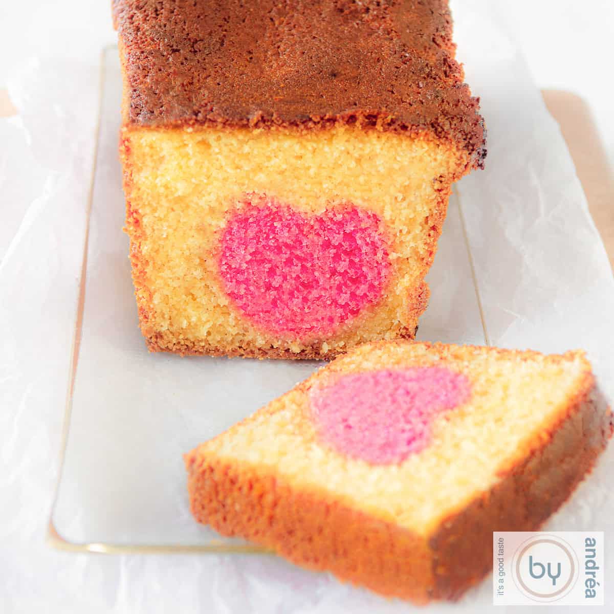 Een vierkante foto met een glazen schaal met daarop een cake waar een plakje is afgesneden. Een roze hart kun je zien