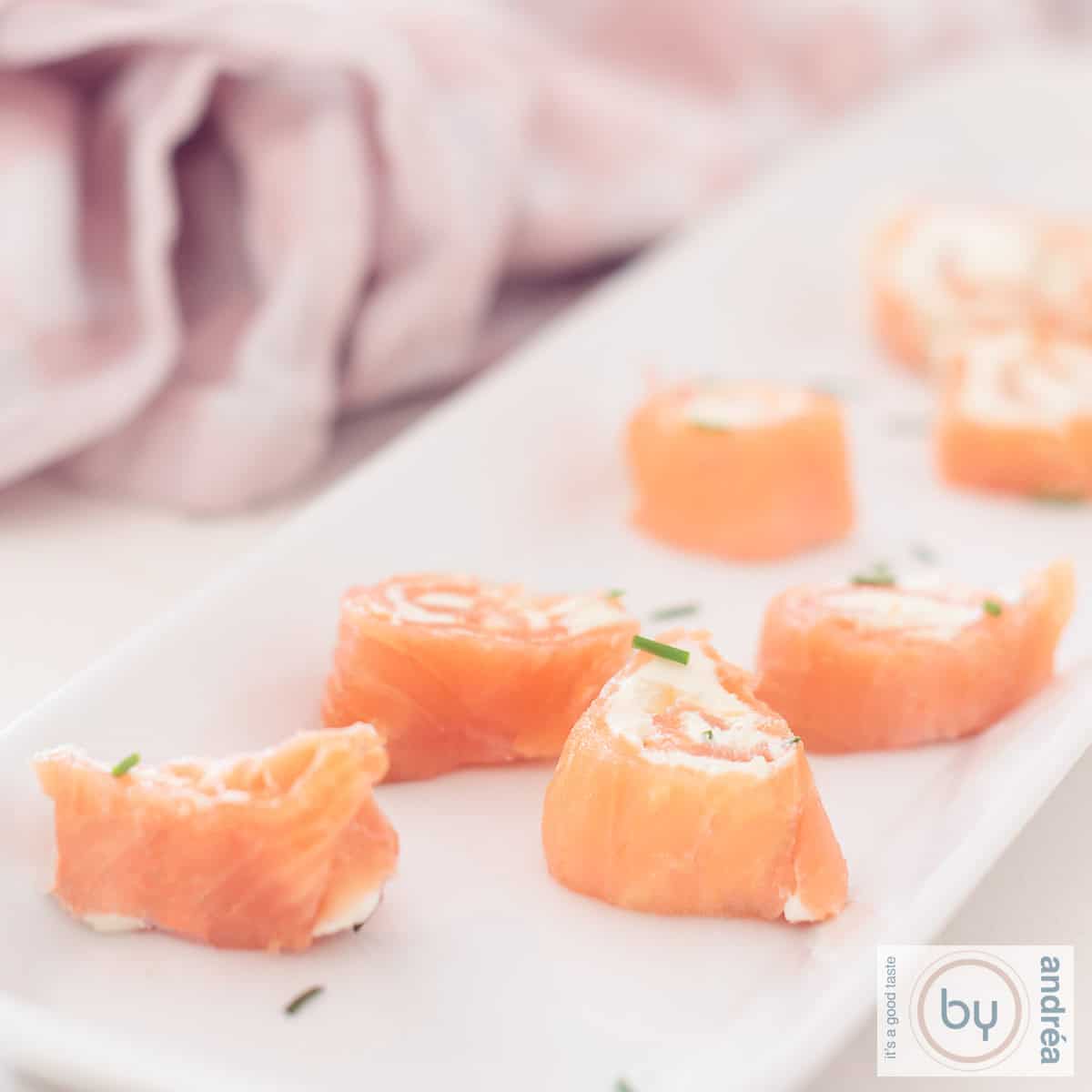 Een vierkante foto met een deel van een lange schaal met rolletjes zalm gevuld met roomkaas en bieslook