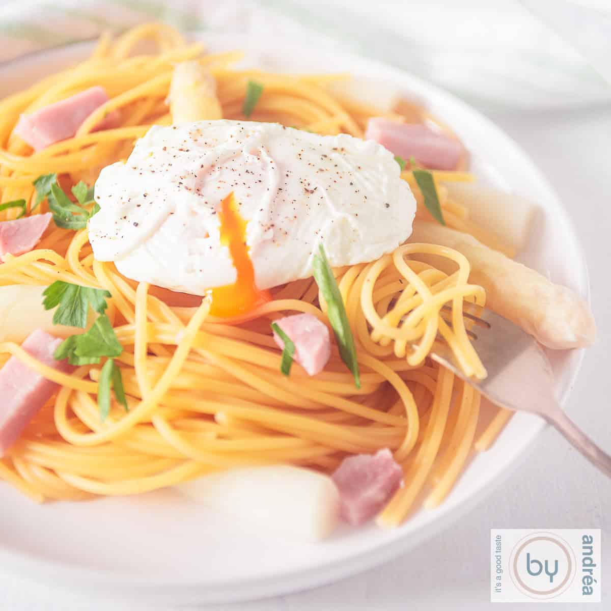 Een vierkante foto van een wit bord met spaghetti, ham, asperges en gepocheerd ei.