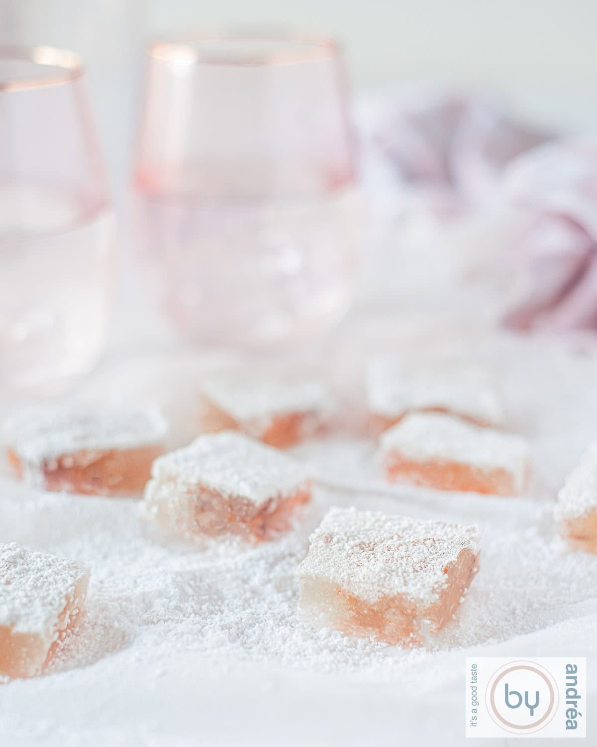 Een witte ondergrond met poedersuiker en daarop blokjes Turks fruit met sinaasappel en walnoot. In de achtergrond twee roze glazen.