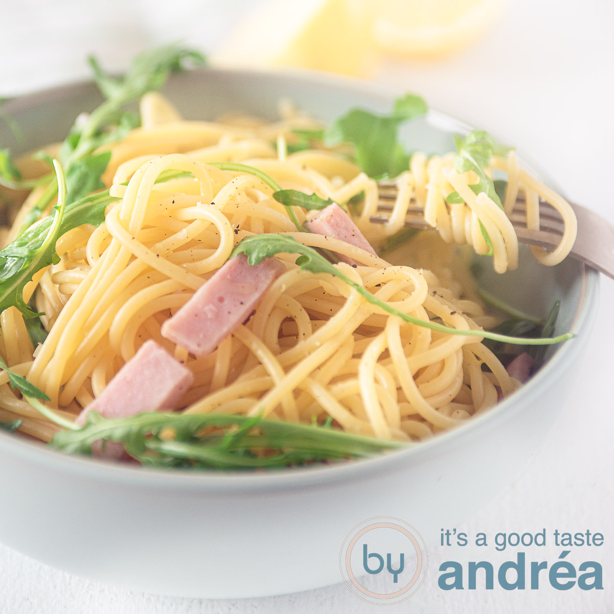 Een vierkante fotonet een grijze schaal gevuld met ham, rucola en spaghetti op een witte ondergrond