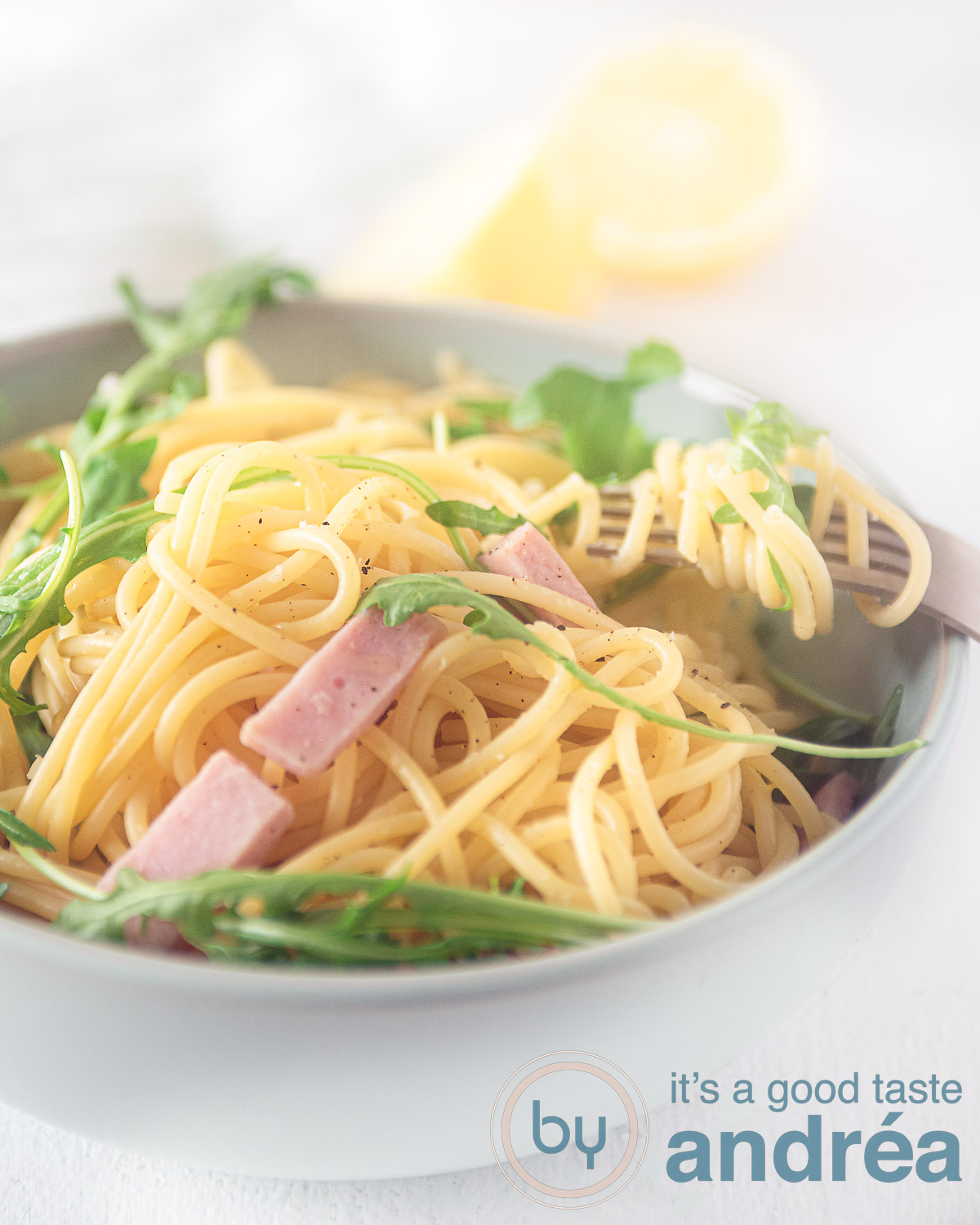 Een grijs schaaltje gevuld met spaghetti, ham, kappertjes en rucola op een witte ondergrond. Bovenin twee halve citroenen.