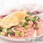 Een vierkante foto met een roze bord in de voorgrond, met een gevulde omelet met rucola, bosui, garnaaltjes en ham. In de achtergrond een roze, witte theedoek.