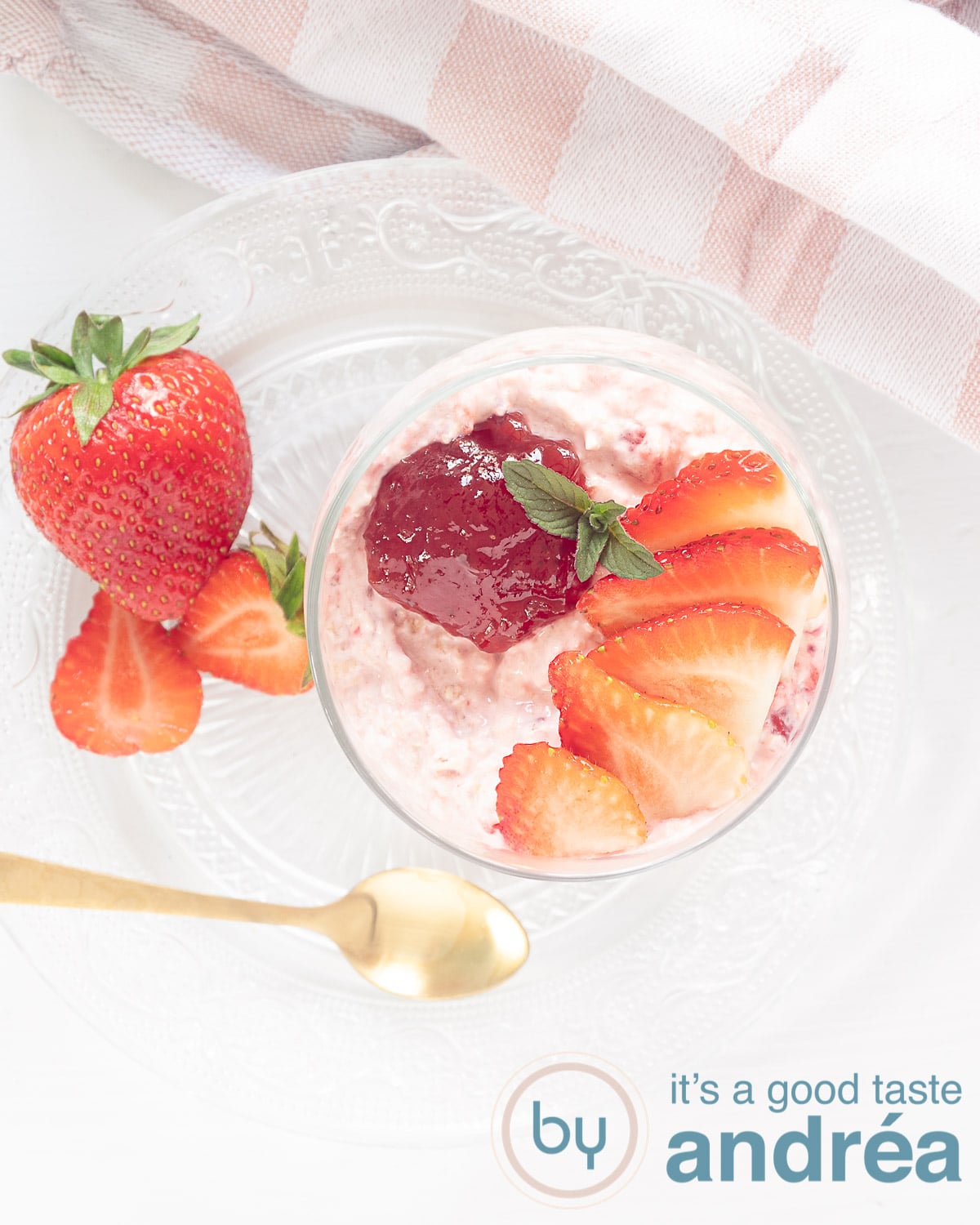 Een foto van bovenaf van een Zomerse Aardbeien smoothie in een kom met een topping van aardbeien en jam. Een witte ondergrond en een gouden lepel
