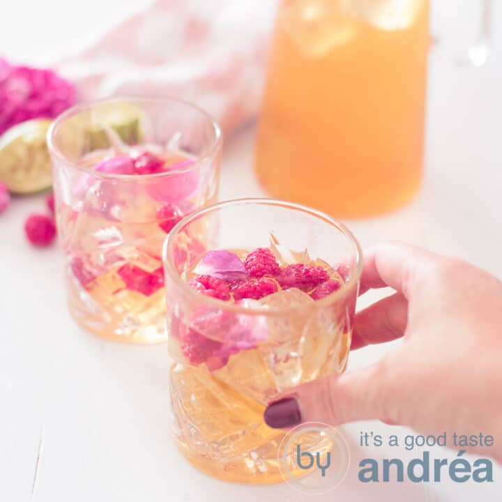 Een vierkante foto met twee glazen ijsthee schuin achterelkaar. Decoratie van roos en framboos en limoen. Een hand pakt het voorste glas.