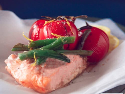 Een vierkante foto van een bakpapier met een zalm met daarop boontjes, gestoofde tomaten en citroenrasp.