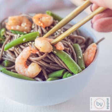 Een vierkante foto met een wit schaaltje gevuld met sobanoodles met garnalen, sperziebonen en sesamzaad. Twee chopsticks pakken een garnaal uit het bakje.