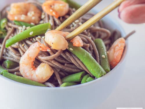 Een vierkante foto met een wit schaaltje gevuld met sobanoodles met garnalen, sperziebonen en sesamzaad. Twee chopsticks pakken een garnaal uit het bakje.
