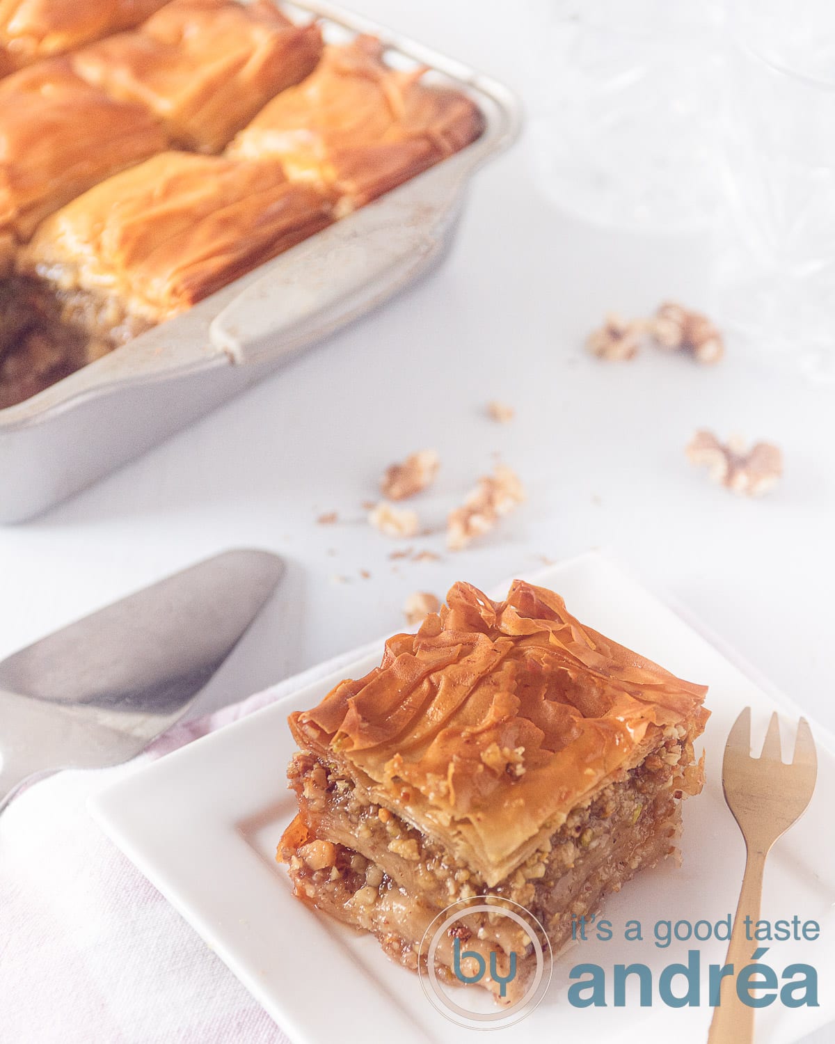 Een foto schuin van boven van een stukje Griekse Baklava op een wit bord met een gouden vork en in de linkerachtergrond een schaal met meer baklava