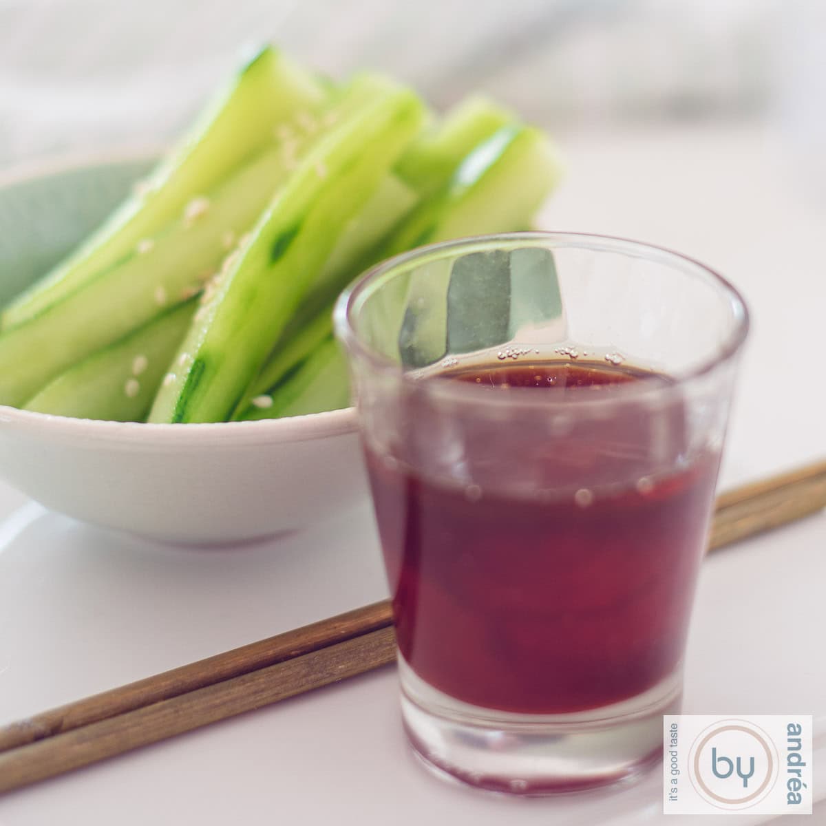 Een vierkante foto met in de achtergrond een schaaltje met reepjes komkommer en sesam, twee eetstokjes en vooraan een glas met een kruidige dipsaus.