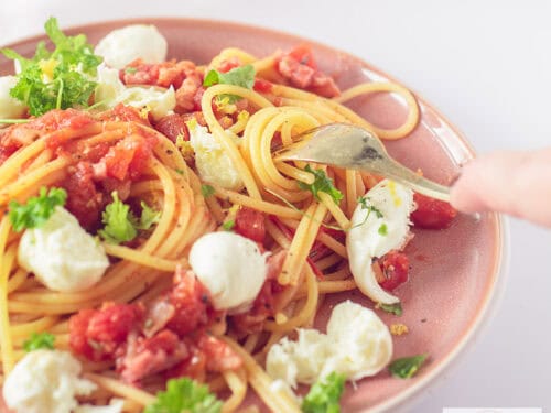 Een vierkante foto van een roze bord gevuld met spaghetti, bacon, tomaten, mozzarella en peterselie