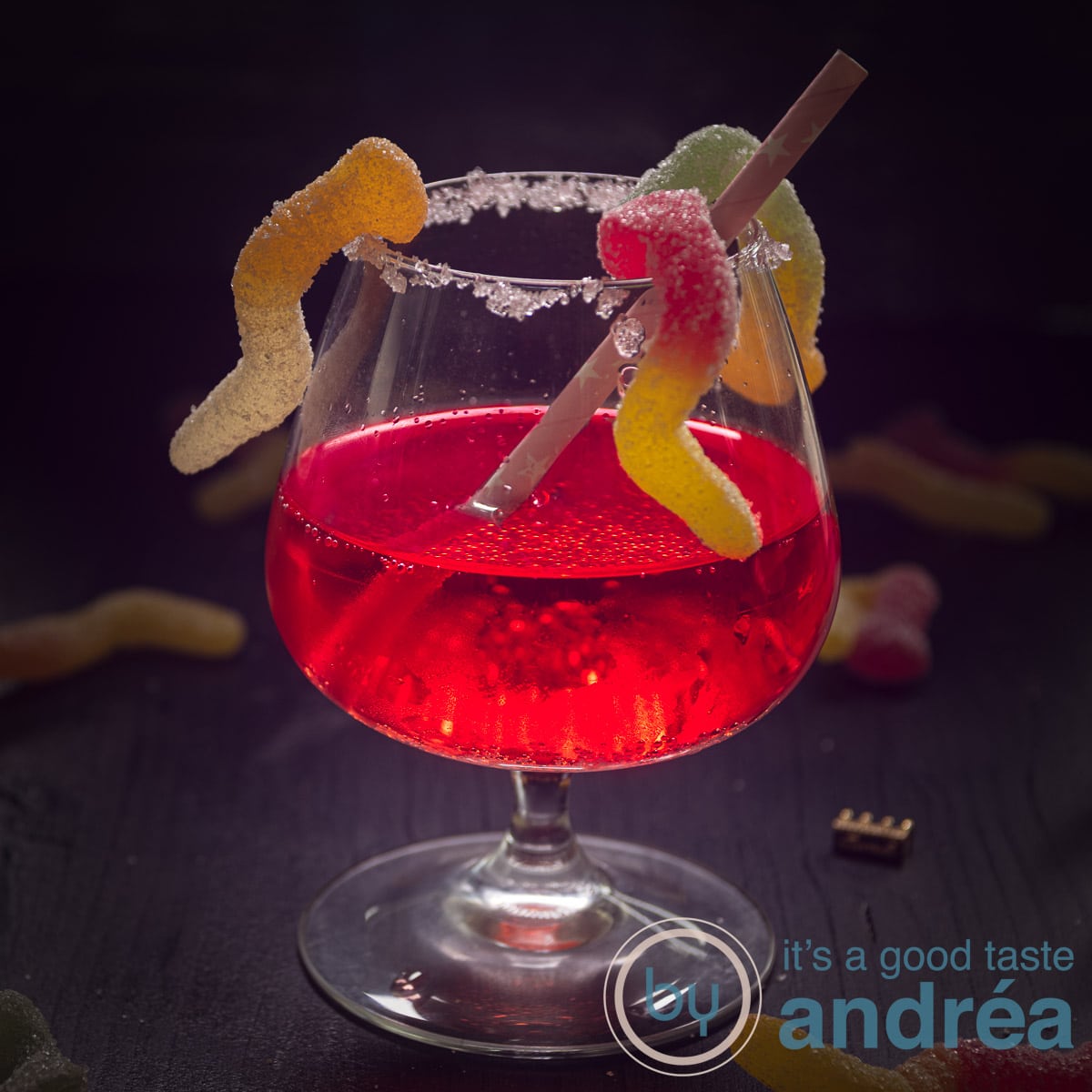 A square photo with a cocktail in the center with Passoa on a black background. Candy worms climb in it.