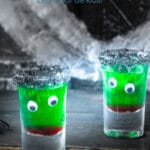 Twee glazen gevuld met een laagje rode siroop en vervolgens groene siroop en een zwart suikerlaagje. Twee oogje erop zodat het een gezichtje lijkt. Een zwarte achtergrond. Een tekst overlay Halloween cocktail zonder alcohol.