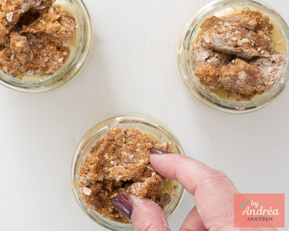 Een hand verdeelt havermoutkruimeldeeg over de appelvulling in drie glazen potjes.