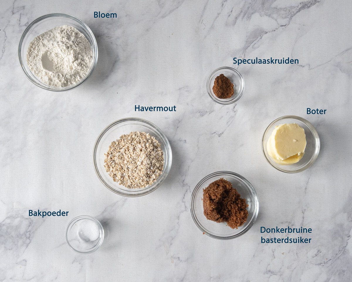Bloem, havermout, speculaaskruiden, boter, donkerbruine basterdsuiker en bakpoeder in schaaltjes met Nederlandse labels.