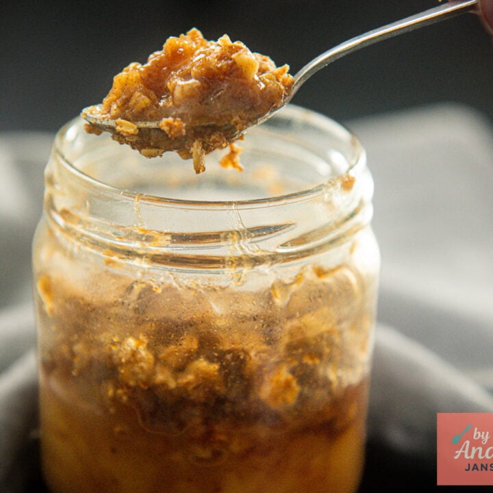 Een hand houdt een lepel met appelstukjes en havermoutcrumble boven een glazen potje appelkruimeltaart.