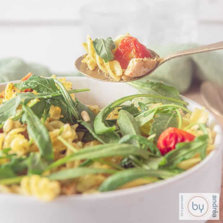 Een vierkante foto met een deel van een witte kom gevuld met fusilli, rucola, tomaten en pesto.