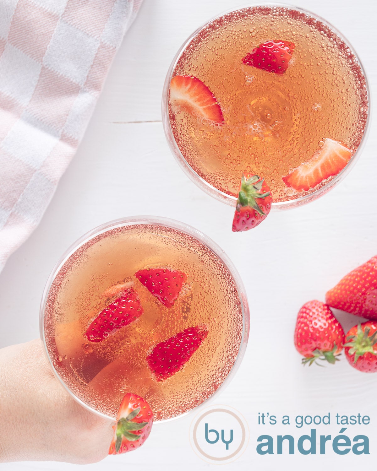 Een foto van bovenaf met twee champagne bowls gevuld met verse aardbeien en Prosecco op een witte ondergrond. Een hand pakt een glas