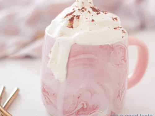 Een vierkante foto van een roze mok met Baileys met warme chocolademelk en een slagroomtopping. Een witte ondergrond, in de achtergrond een roze witte theedoek. Links voorin twee gouden rietjes