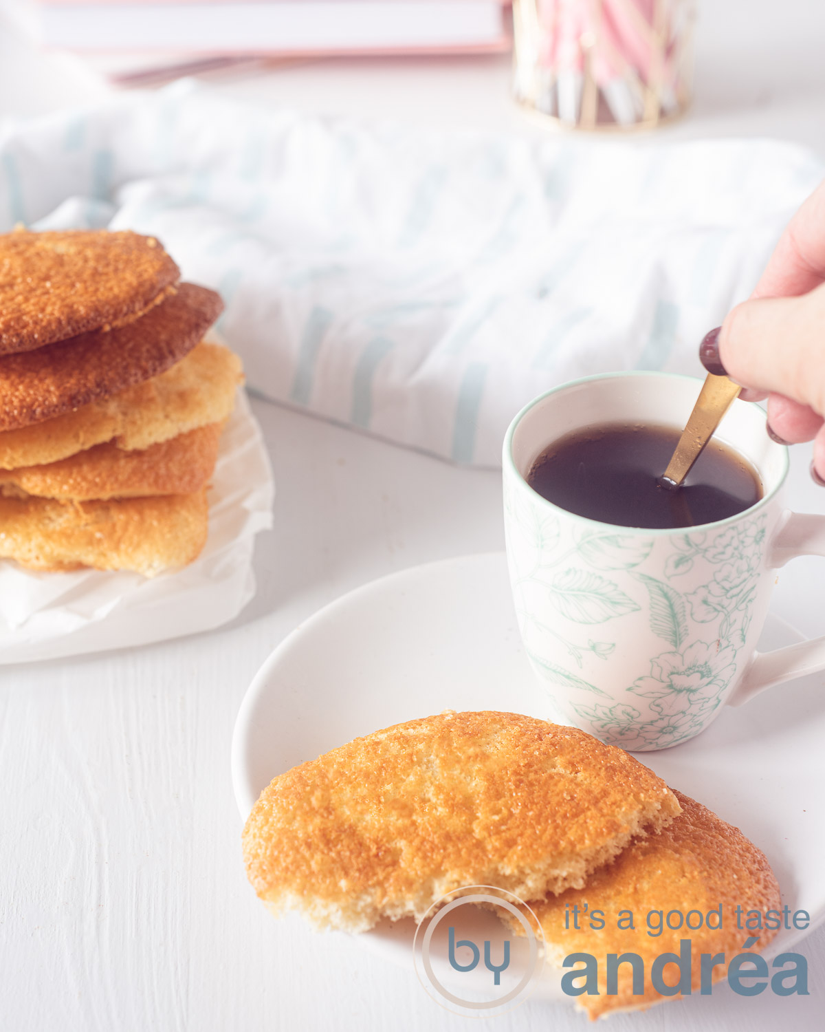 Een wit bord met twee halve eierkoeken en een kopje koffie (waar de lepel wordt geroerd. Aan de linkerkant een stapel eierkoeken. Een witte ondergrond.