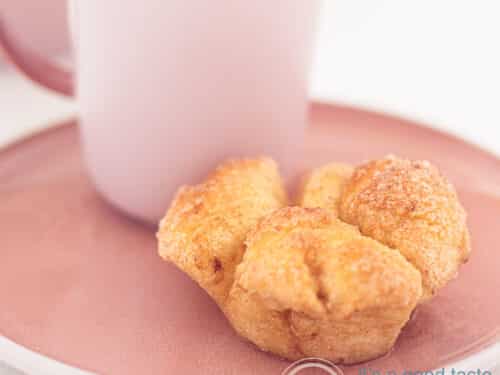 Een vierkante foto van een roze bord met daarop een kopje koffie en daarvoor een monkey bread muffin.