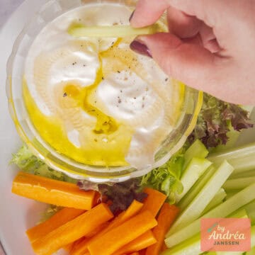 Een vierkante foto met romig geklopte feta dip met olijfolie en groente sticks eromheen. Een hand dipt groente in de feta saus.