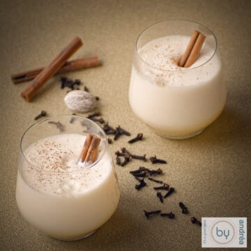Een vierkante foto met twee glazen gevuld met traditionele eggnog met een kaneelstokje. er tussen allemaal kruiden die je nodig hebt.