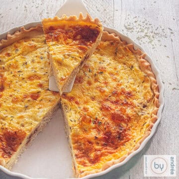 Een vierkante foto van een grijze schaal gevuld met geitenkaas sjalot quiche. Een punt is eruit gesneden en ligtop een wit taarschep erop.