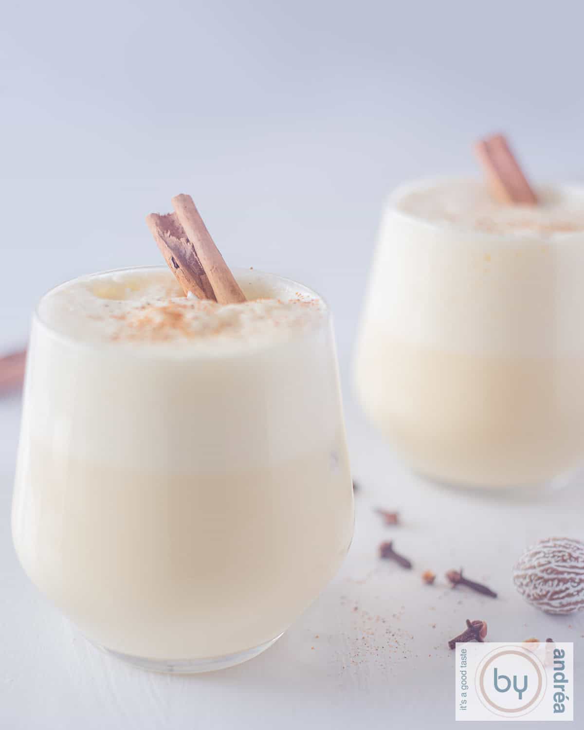 Een hoogtefoto met twee glazen gevuld met eggnog en een kaneelstokje. Kruiden die liggen ertussen in. Een witte ondergrond