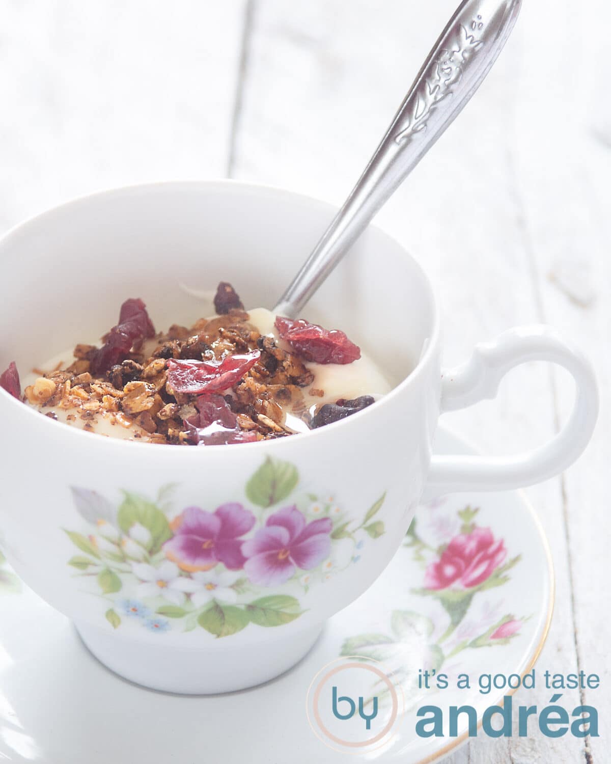 Een kopje gevuld met yoghurt en muesli uit de oven met cranberries. Een zilveren lepel steekt erin.