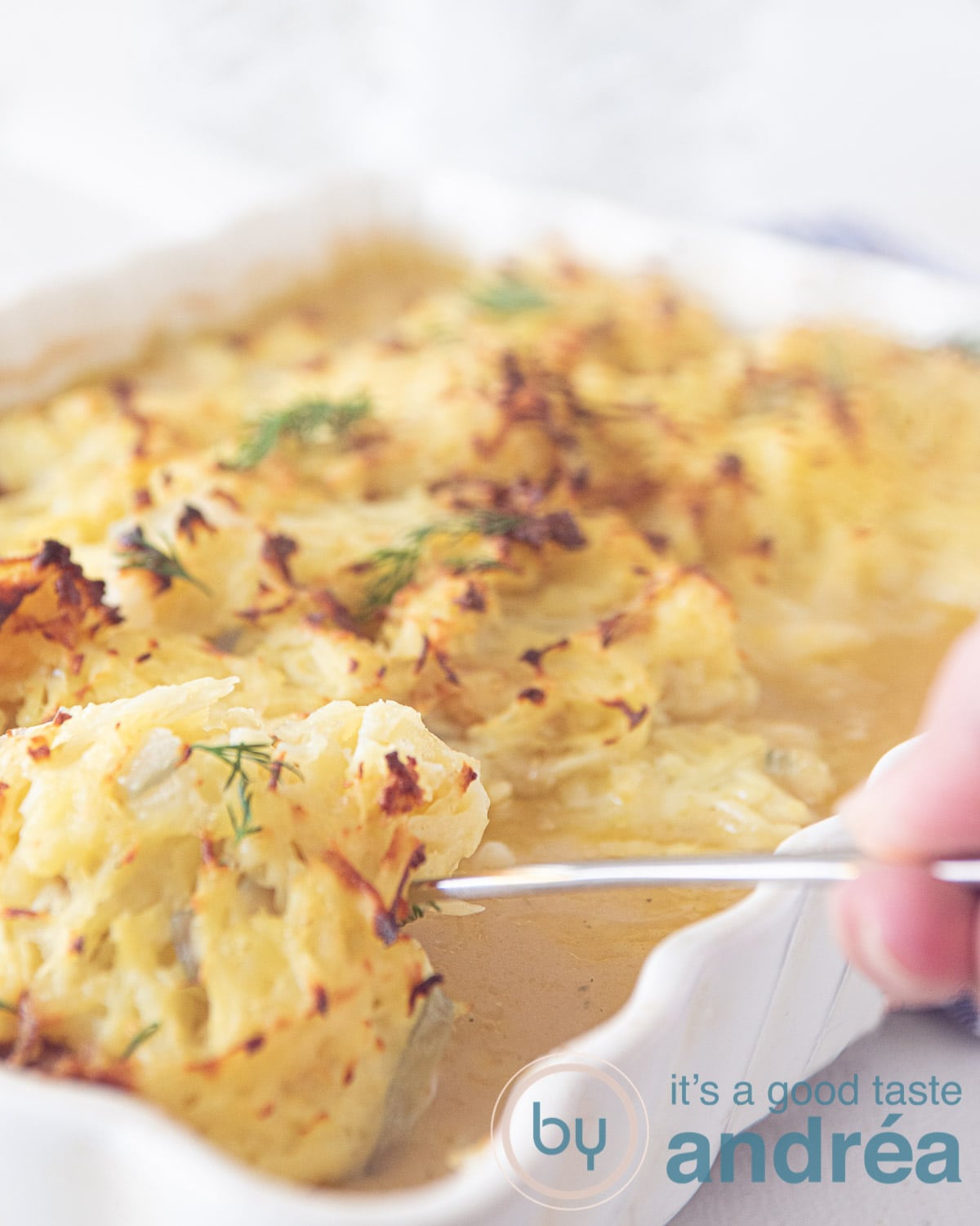 Een witte ovenschaal waarbij een hand een kabeljauw met rösti topping uit de schaal schept