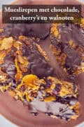 Repen op een roze bord met chocola. Een tekst bovenin: mueslirepen. met chocolade, cranberry's en walnoten