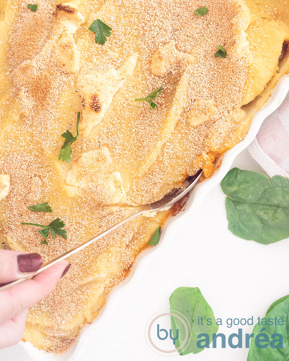 A photo from above of a white baking dish with a gratin. A hand puts a spoon in it. Spinach leaves bottom left