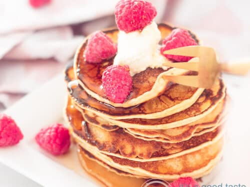 Een vierkante foto met een stapel pancakes, frambozen erop en omheen en een dot crème fraîche. In de achtergrond een roze witte doek