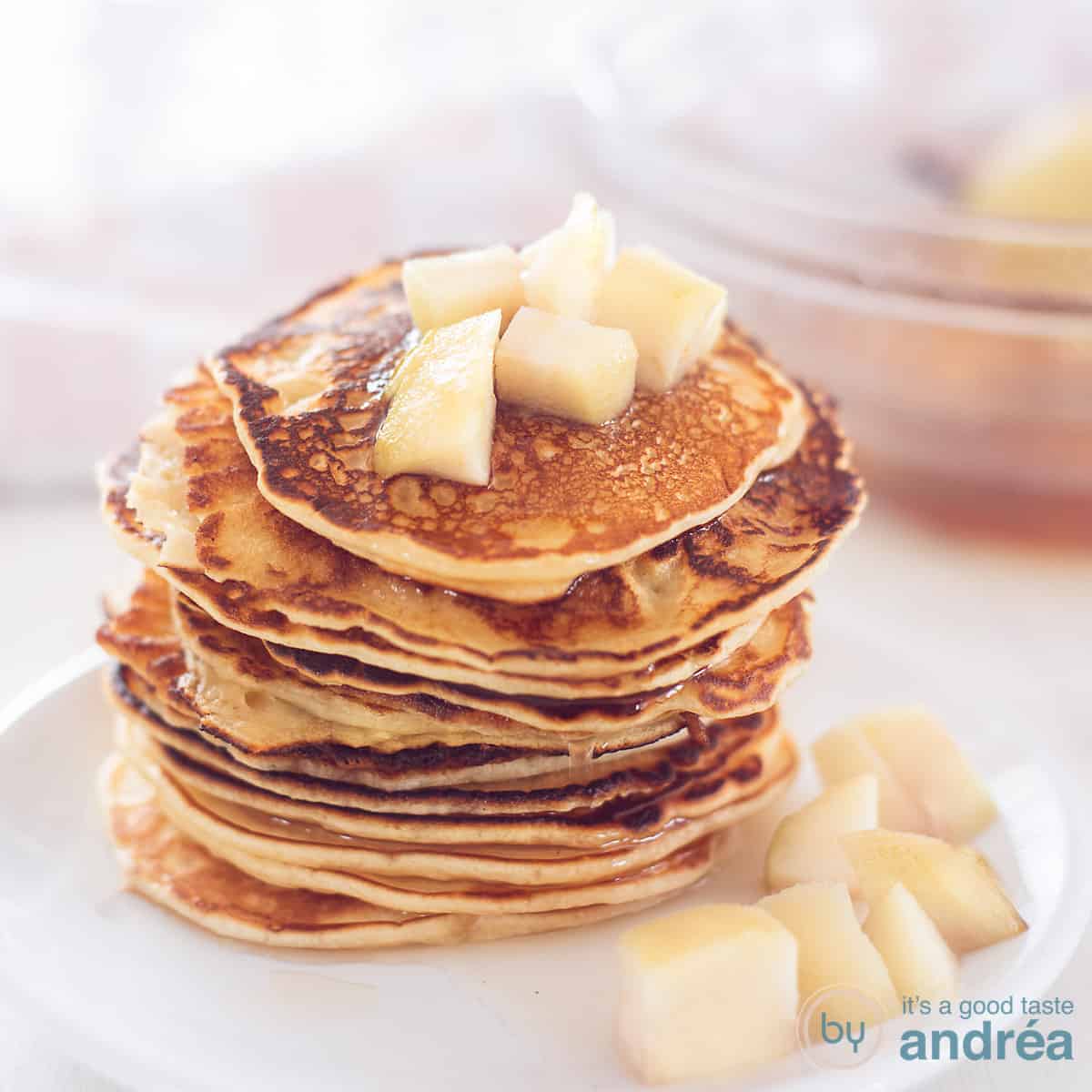 Een vierkante foto met een wit bord met een stapel pancakes en daarop en omheen stoofperen.
