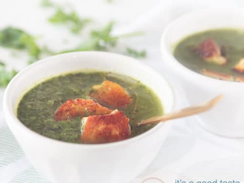 vierkante foto met twee witte kommen met soep van spinazie, boerenkool en paksoi. Met een topping van Halloumi