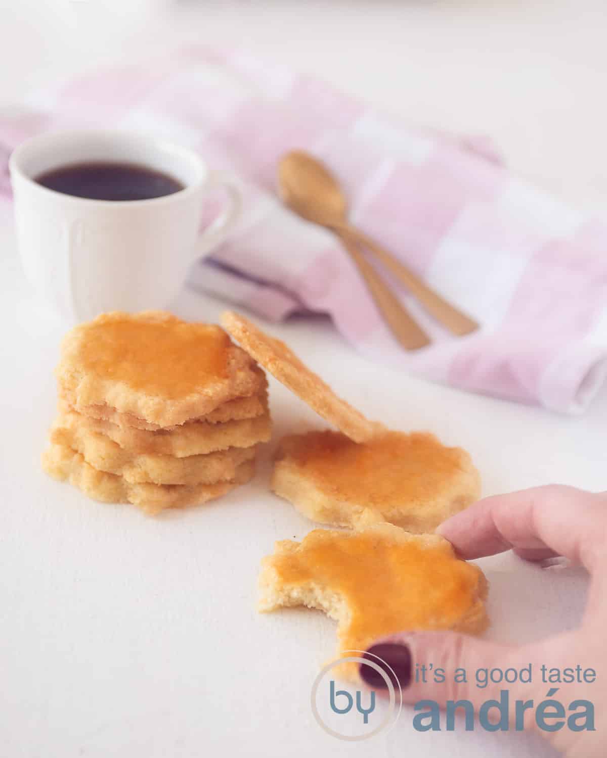 Vijf sabelkoekjes op elkaar. Twee aan de zijkant. Vooraan een koekje met een hapje. Een hand pakt dit koekje. Achteraan een witroze handdoek en een kopje koffie.