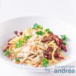 Een vierkante foto met een diep wit bord gevuld met pasta, pesto van boerenkool, geraspte kaas en zongedroogde tomaten. Gegarneerd met peterselie