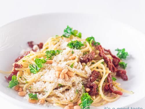 Een vierkante foto met een diep wit bord gevuld met pasta, pesto van boerenkool, geraspte kaas en zongedroogde tomaten. Gegarneerd met peterselie