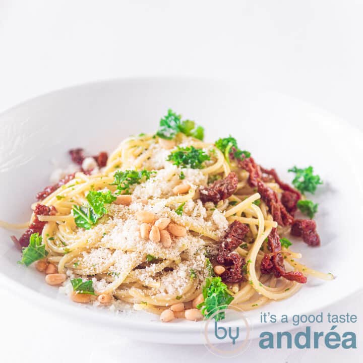 Een vierkante foto met een diep wit bord gevuld met pasta, pesto van boerenkool, geraspte kaas en zongedroogde tomaten. Gegarneerd met peterselie