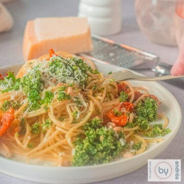 Een vierkante foto met een wit bord met spaghetti, boerenkoolpesto en zongedroogde tomaten. Een vork prikt in het bord.