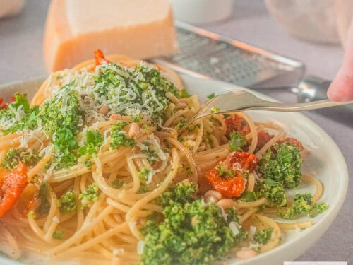 Een vierkante foto met een wit bord met spaghetti, boerenkoolpesto en zongedroogde tomaten. Een vork prikt in het bord.