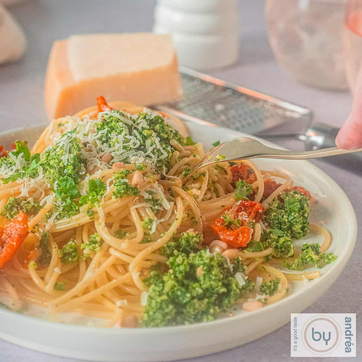 Een vierkante foto met een wit bord met spaghetti, boerenkoolpesto en zongedroogde tomaten. Een vork prikt in het bord.