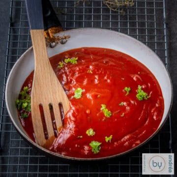 Een vierkante afbeelding met een koekenpan gevuld met enchiladasaus. Een houten lepel in de koekenpan.