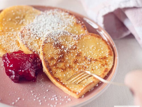 Een vierkante foto met een roze bord met hartvormige pannenkoeken met poedersuiker en aardbeienjam. Een vork prikt in een pannenkoek.