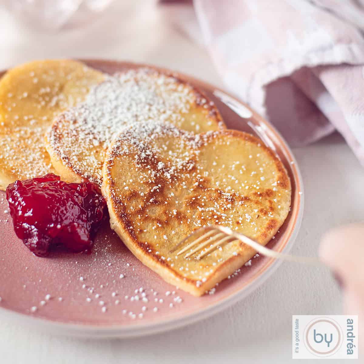 Een vierkante foto met een roze bord met hartvormige pannenkoeken met poedersuiker en aardbeienjam. Een vork prikt in een pannenkoek.