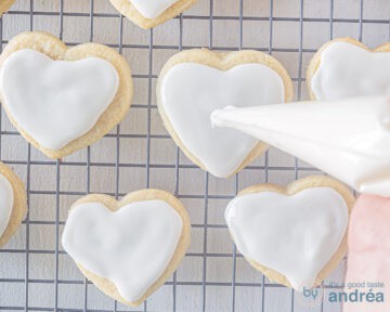 Een rek met daarop hartjeskoekjes. Een spuitzak spuit witte icing op elk koekje