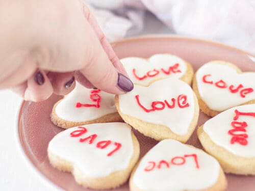 Een vierkante foto met een roze bord met hartjeskoekjes met icing en een tekst in het rood. Een hand pakt een koekje.