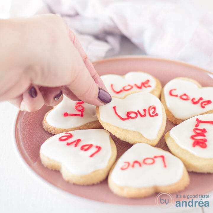 Een vierkante foto met een roze bord met hartjeskoekjes met icing en een tekst in het rood. Een hand pakt een koekje.