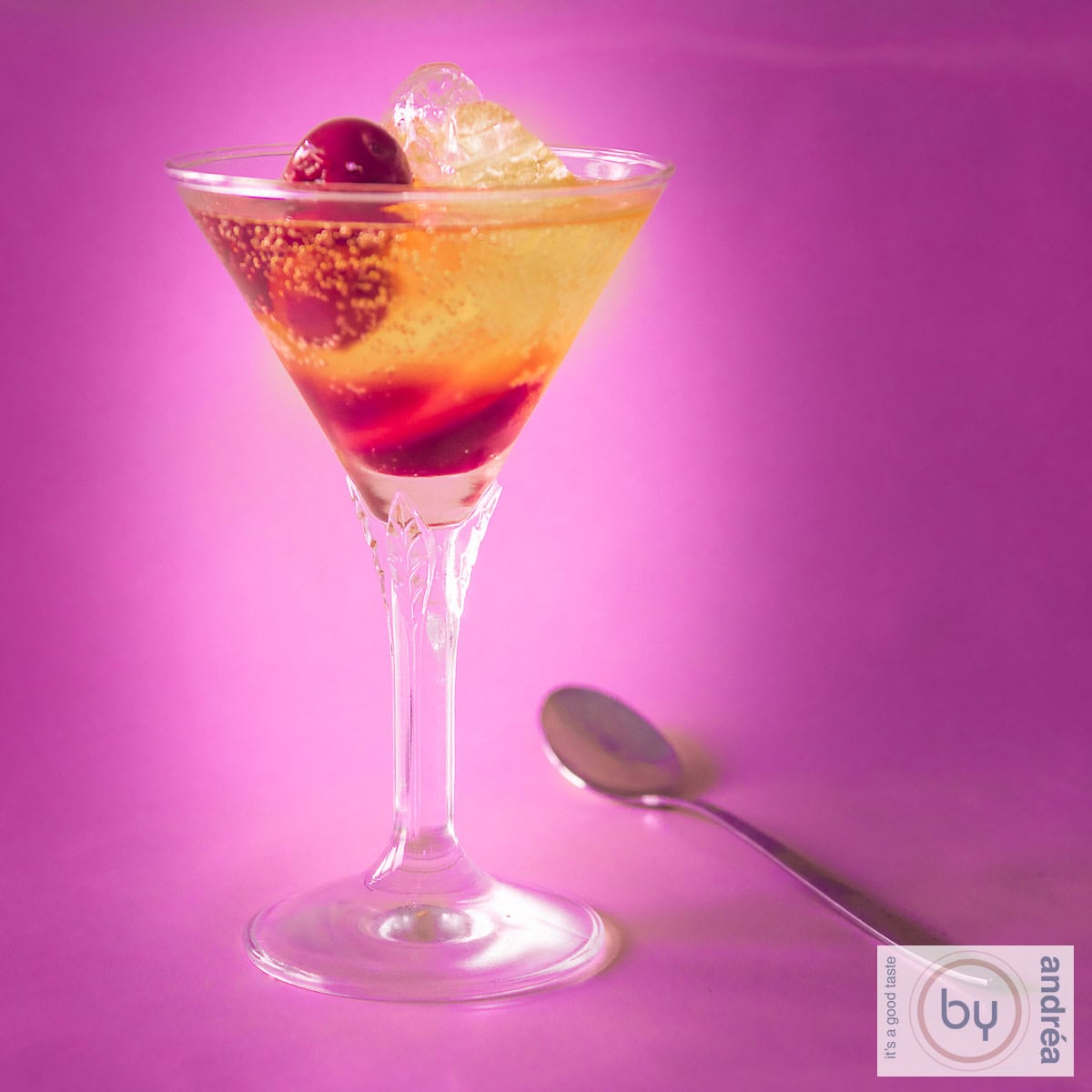 Een vierkante foto met een roze achtergrond. Erop een cocktail glas met een kersen prosecco cocktail. Een gouden lepel ligt er naast.